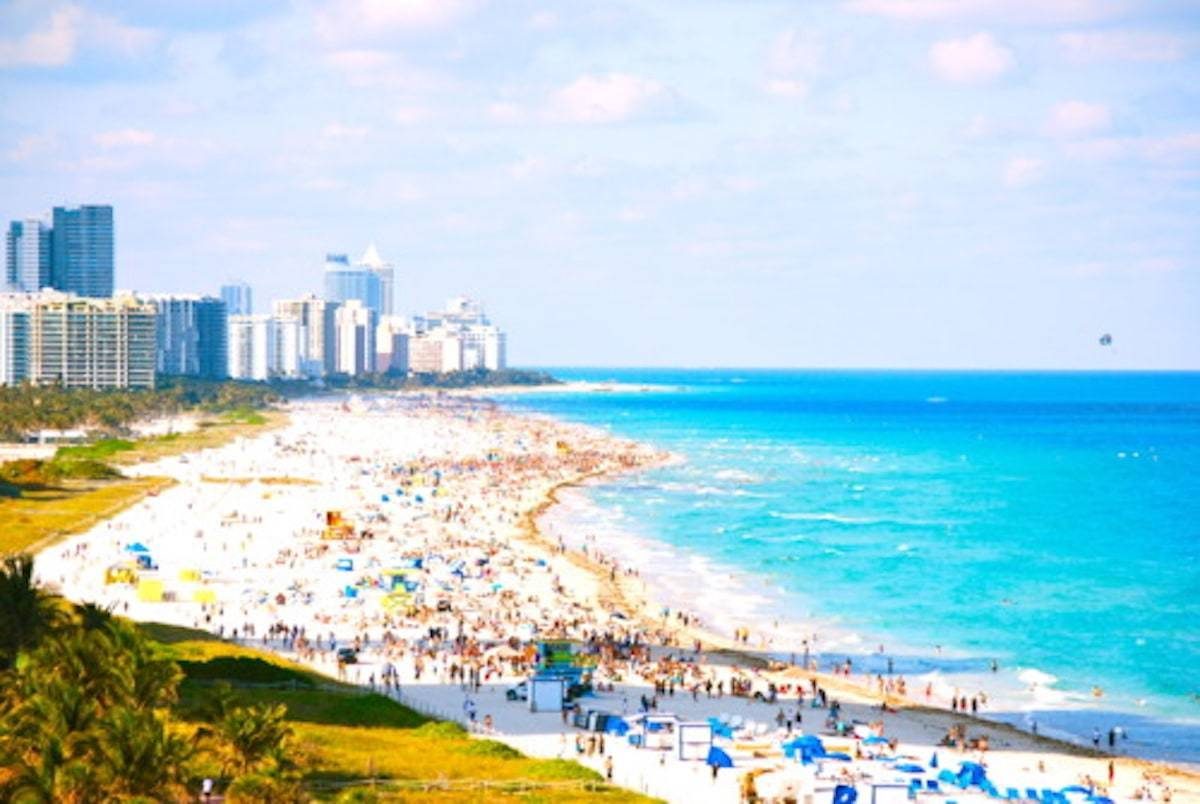 Ocean shore with people filling the sand near various buildings.